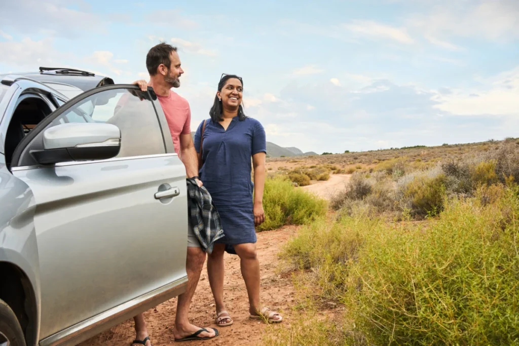 A couple standing next to an SUV in a scenic outdoor setting, with the man leaning against the car door and the woman looking into the distance, both smiling.