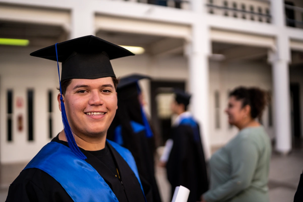 Image of a smiling graduate wearing a black cap with a blue tassel and a blue graduation stole over a black gown. He is standing in front of a building, with other graduates visible in the background.