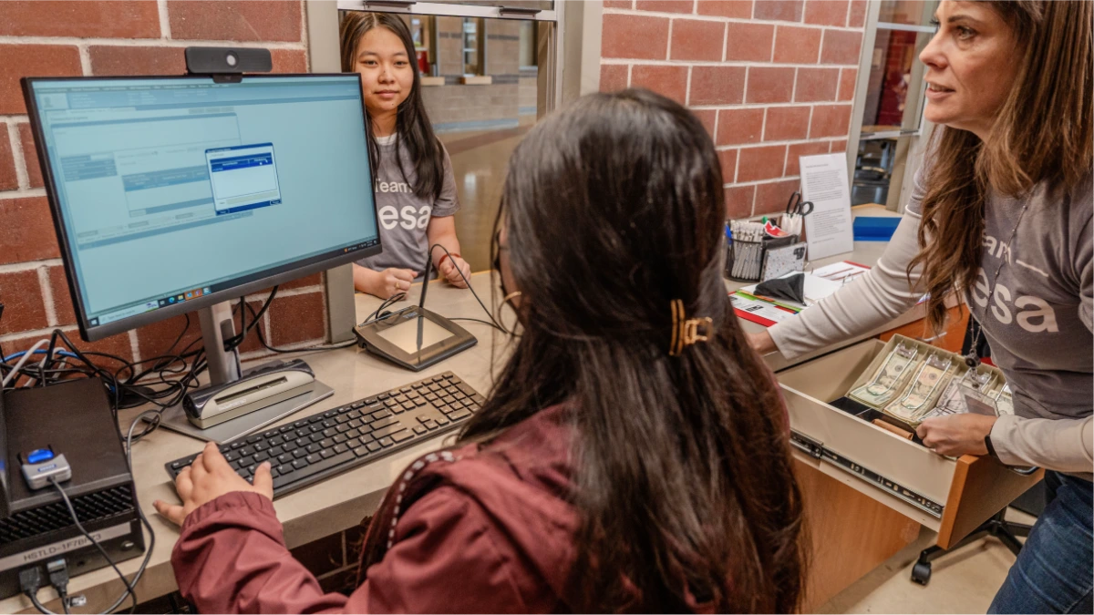 Three women at a desk inside a High School Credit Union, one woman typing at a computer while others assist, with cash visible on the desk