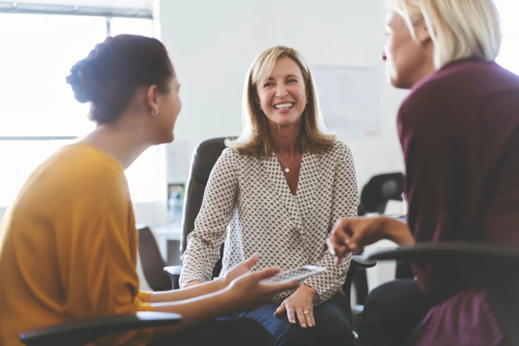 The image shows three women sitting together in a casual office environment, engaged in a friendly conversation. One woman, in the center, is smiling warmly, while the other two women face her, listening and chatting. The atmosphere is light and collaborative, with the women appearing relaxed and engaged in their discussion.