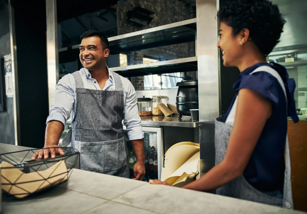 The image shows two people working in a restaurant or cafe setting. Both are wearing aprons and appear to be engaged in a light conversation. The man on the left is smiling warmly while resting his hand on the counter, and the woman on the right is turned slightly towards him, also smiling. The background reveals part of a kitchen with various cooking equipment and ingredients, indicating a friendly and relaxed work environment.