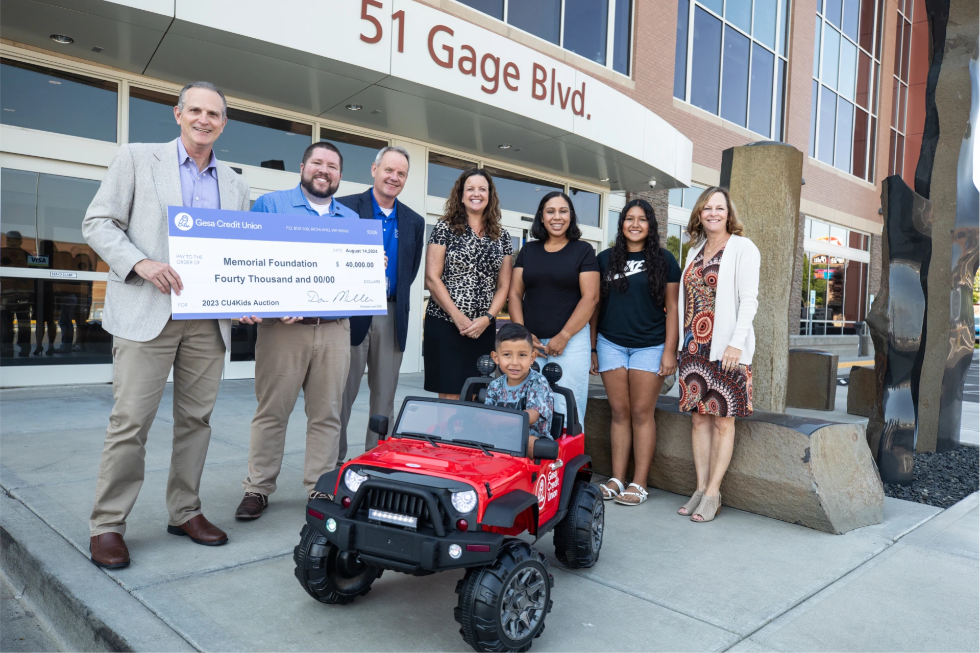 Group of people standing outside 51 Gage Blvd., holding a large check for $40,000 donated by Gesa Credit Union to the Memorial Foundation, with a young child sitting in a toy red jeep bearing the credit union's logo.