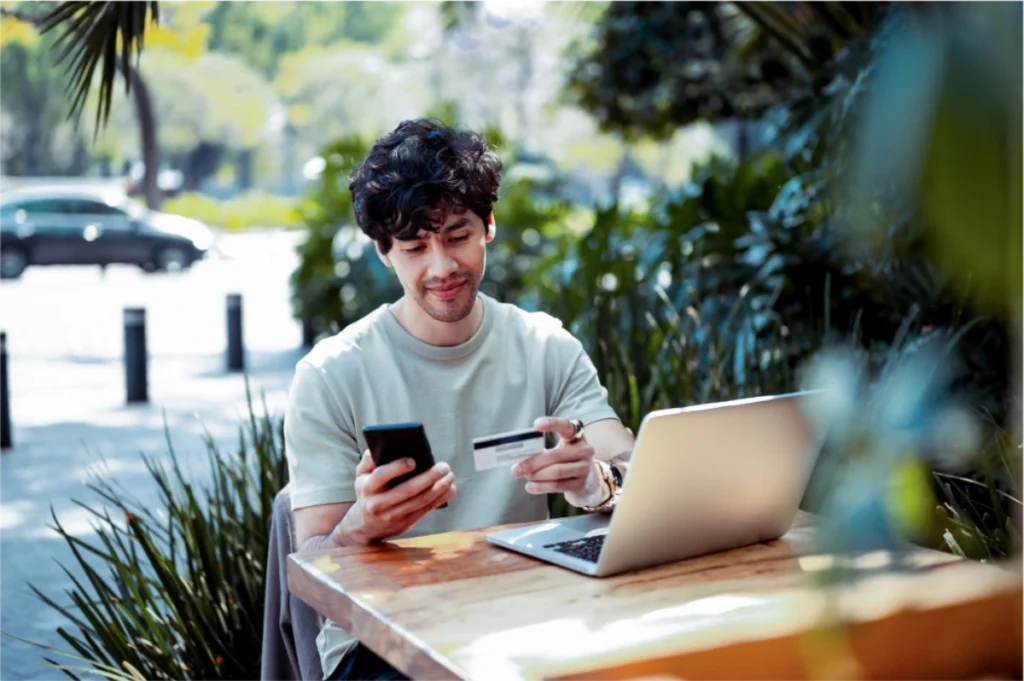 Young man sitting outdoors at a wooden table, holding a smartphone and credit card while using a laptop, surrounded by greenery and a city street in the background.