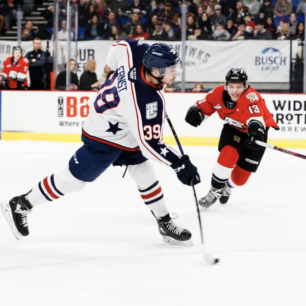 Two ice hockey players in action during a game. The player in the foreground, wearing a white jersey with the number 39 and a blue helmet, is taking a shot with his hockey stick. Another player in a red and black jersey, wearing the number 13, is in pursuit from behind. The background shows spectators and boards with advertisements.