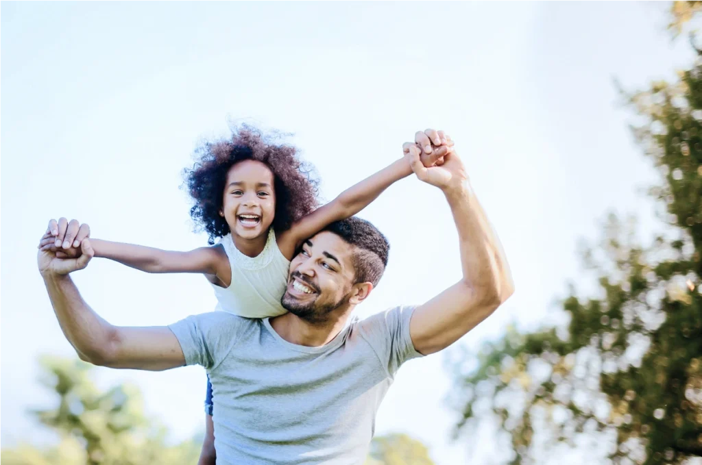 A joyful moment between a father and daughter - he carries her on his shoulders outdoors against a bright sky while both laugh, the young girl's curly hair flowing as she spreads her arms wide with happiness