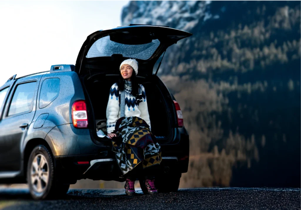 A young woman in winter clothing - a white knit hat, patterned sweater, and geometric print pants - sits in the open trunk of an SUV against a mountainous backdrop.