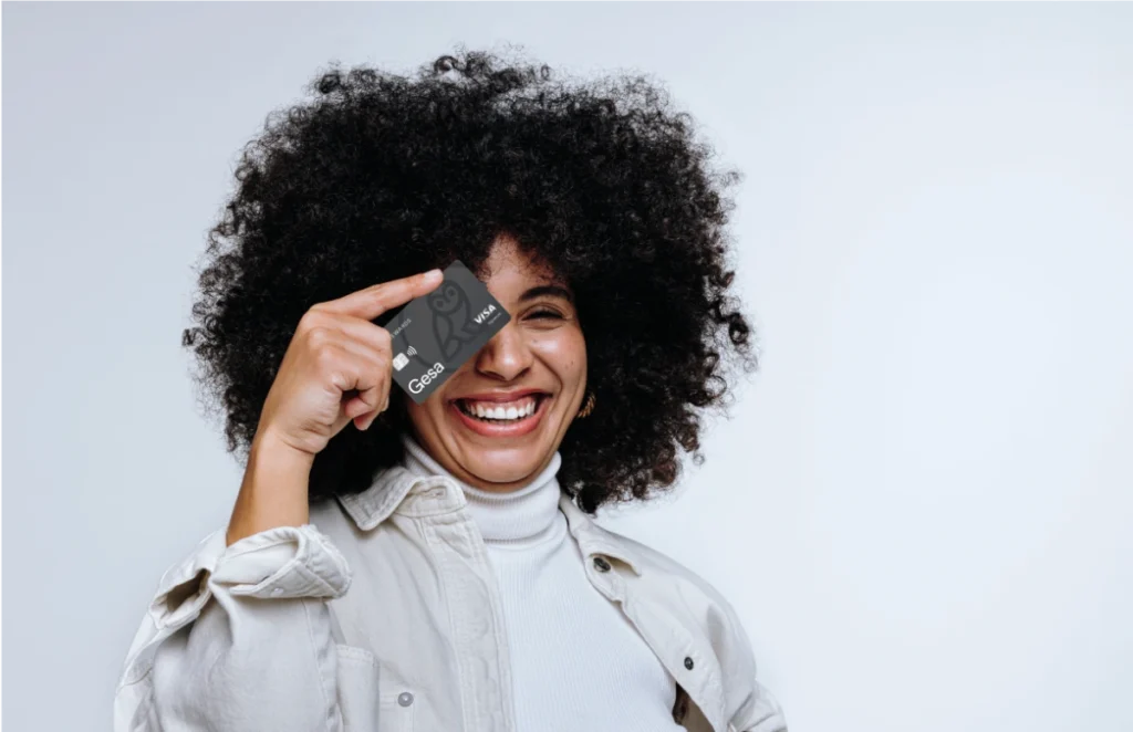 A woman with a large natural afro hairstyle smiles broadly while holding a Gesa credit card over one eye. She's wearing a white turtleneck and light colored jacket against a white background