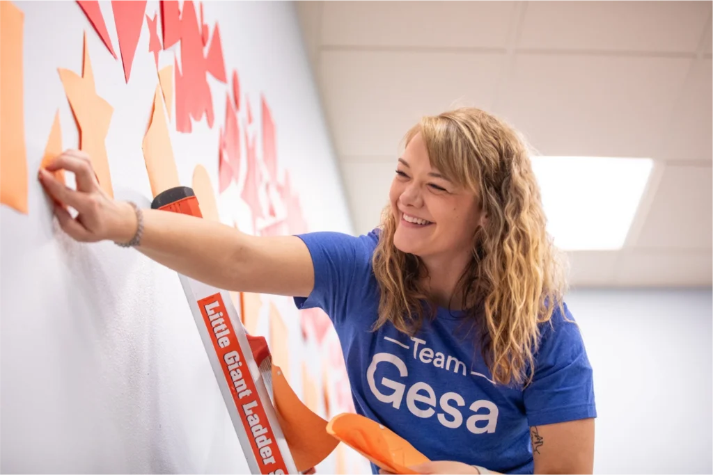 A smiling woman wearing a 'Team Gesa' shirt decorates a wall with orange paper shapes while standing near a ladder indoors.